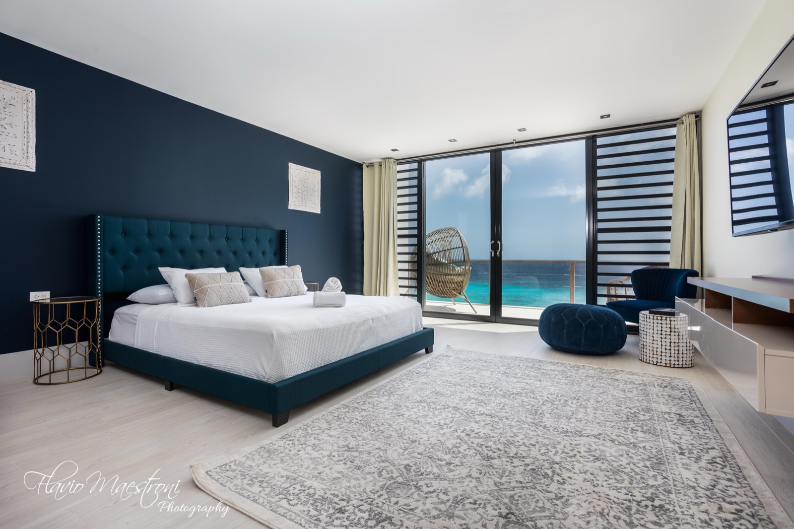 Luxury Bonaire villa master suite with a plush king bed, deep blue accent wall, and a private balcony with a hanging chair overlooking the turquoise Caribbean Sea.